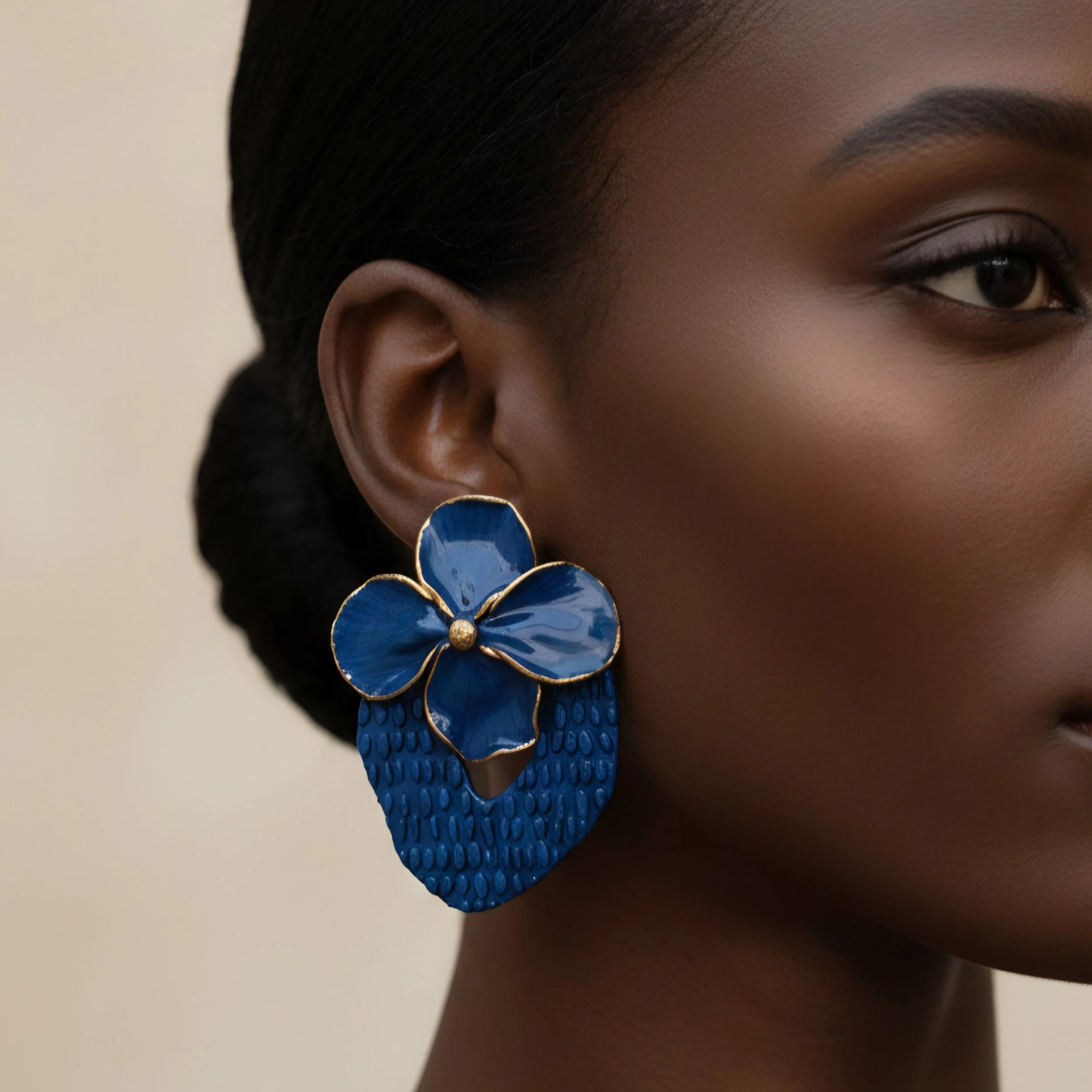 Un gros plan d'une partie du visage d'une femme noire, montrant l'œil, la partie de l'oreille, un boucle d'oreille dorée, une collier bleu en forme de fleur et un fond beige.