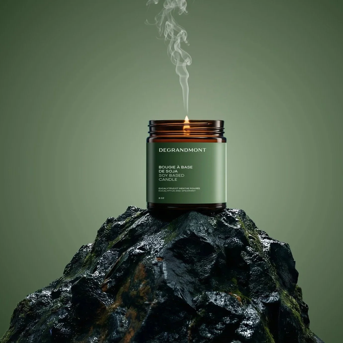 Une bougie parfumée à base de soja avec une étiquette verte, posée sur une roche noire humide, avec de la vapeur s'élevant de la flamme contre un fond vert. La bougie est de la marque Degrandmont, avec un parfum d'eucalyptus et de menthe poivrée.