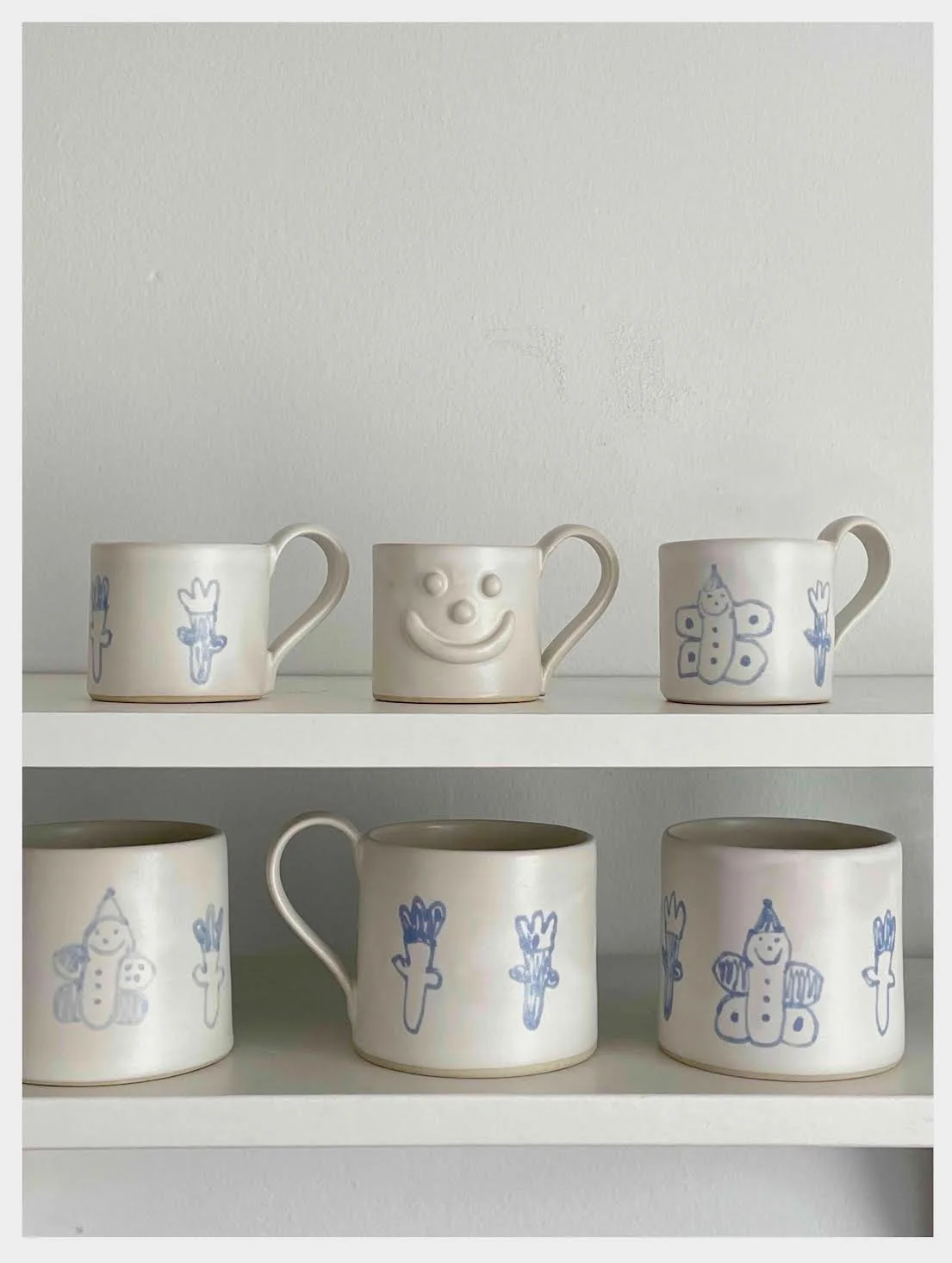 Série de six tasses en porcelaine, décorées de dessins de dessins d'enfants, avec différentes figures comme un soleil, une poupée, un papillon, et des carottes dessinées à la main, exposées sur une étagère blanche.
