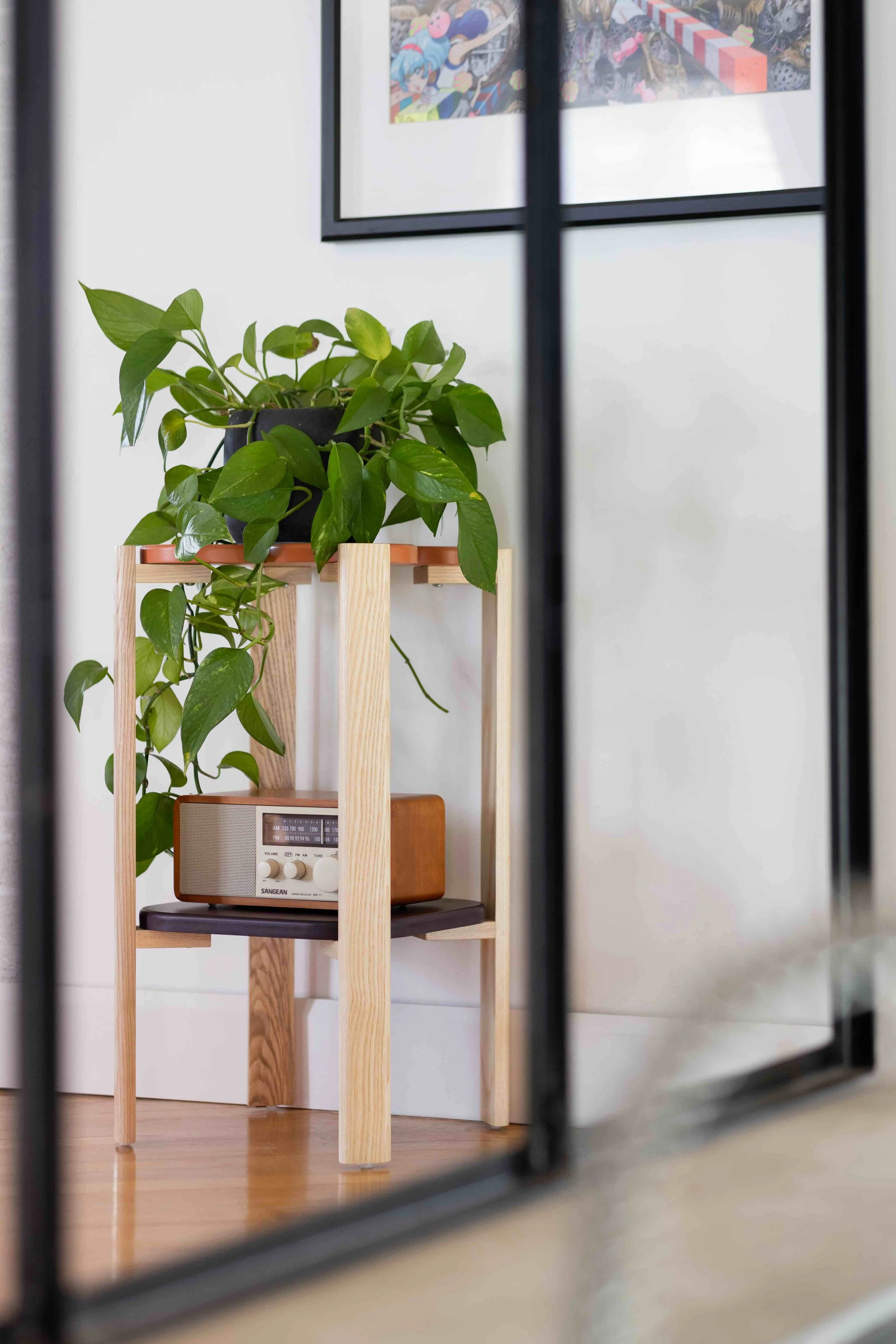 Une plante verte en pot sur une étagère en bois, avec une radio rétro en dessous, dans une pièce lumineuse avec un tableau d'art coloré au mur.