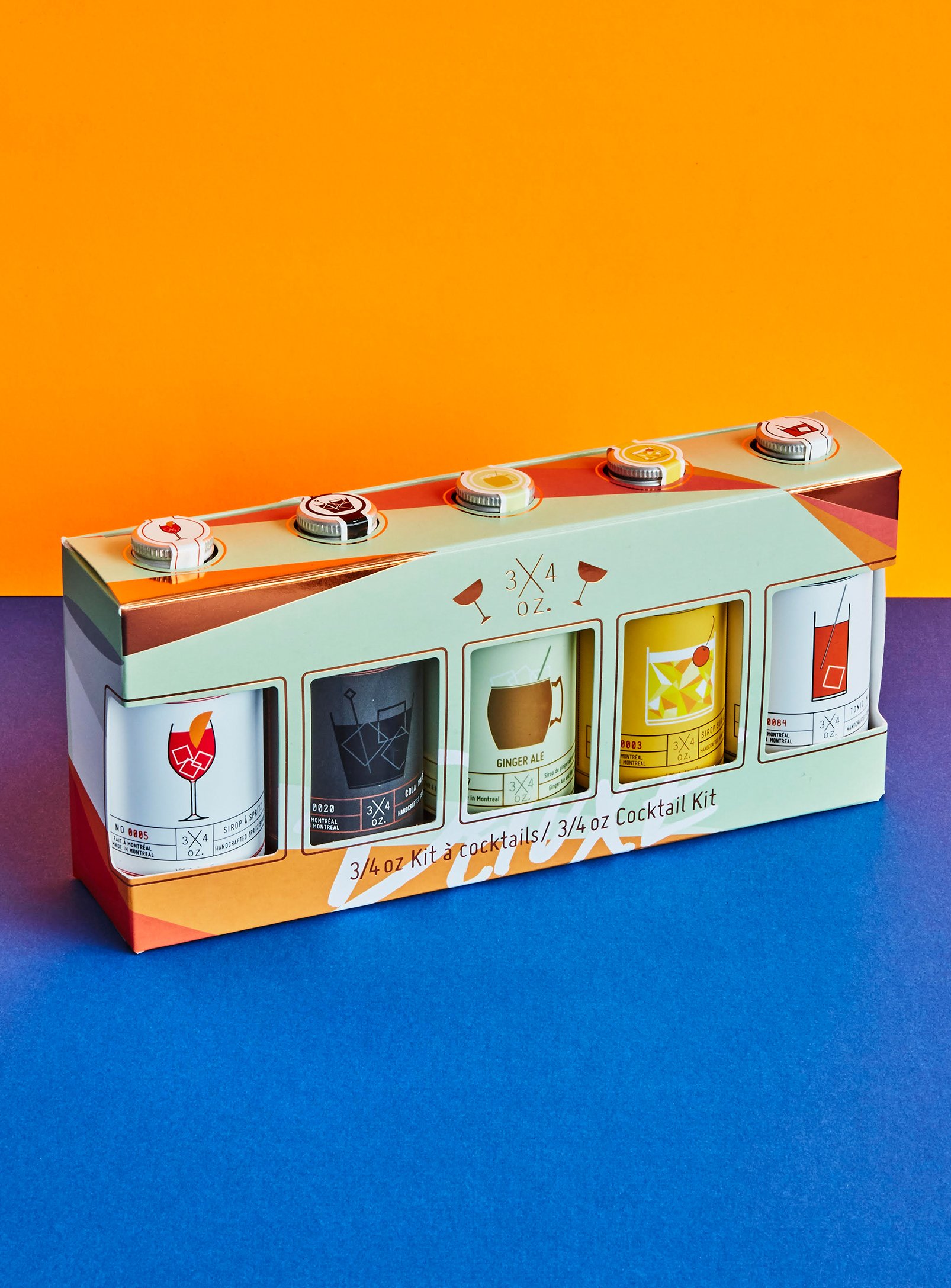 Une boîte de kit à cocktails contenant quatre petites bouteilles avec différentes recettes sur la boîte, dont une Margarita, un Ginger Ale, un Cocktail mystérieux et un autre cocktail dont l'image n'est pas entièrement visible, sur un fond coloré avec un mur orange et une surface violette.