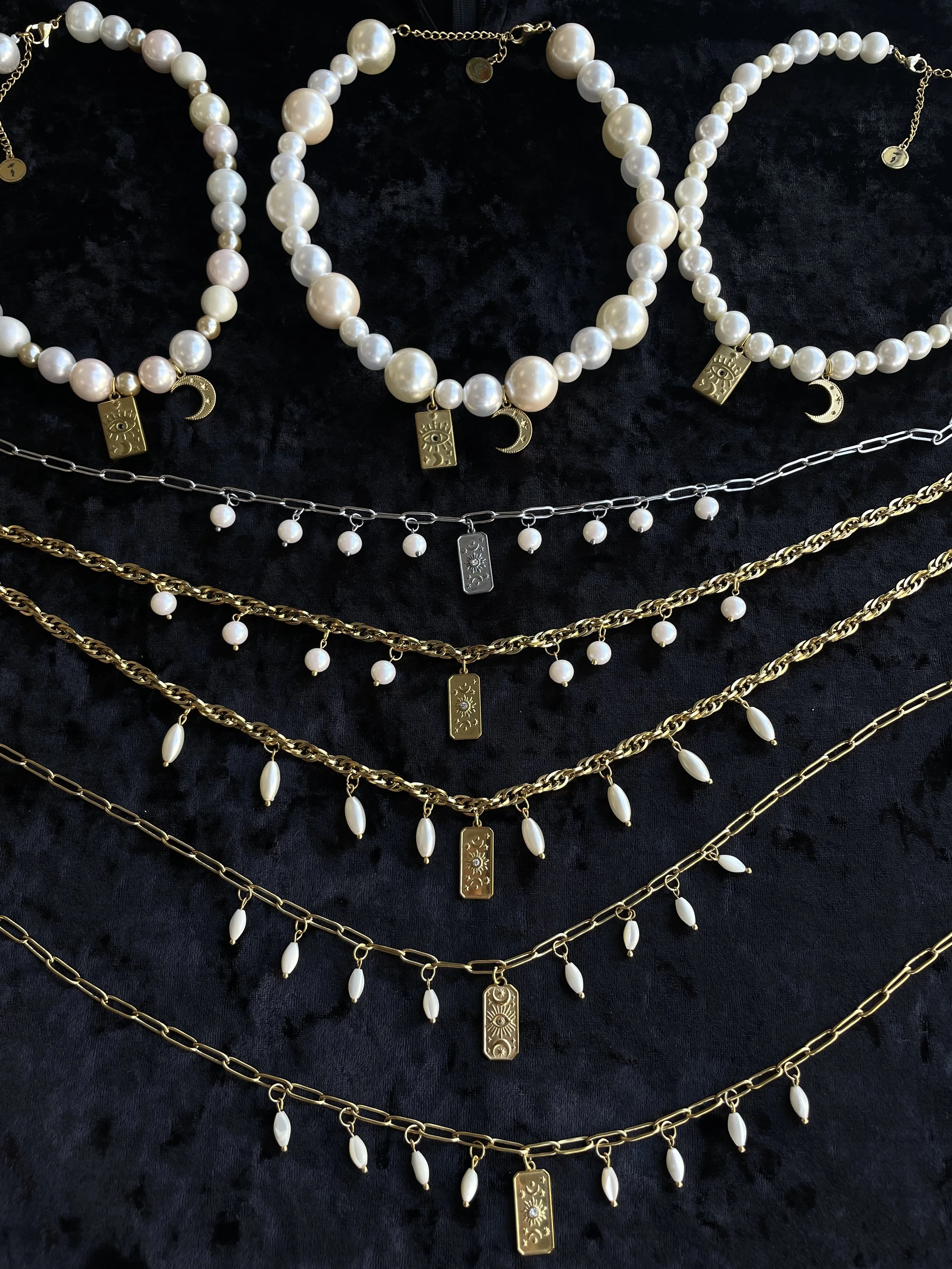 Collection de bijoux en perles blanches et chaînes en or et argent, comprenant des colliers avec pendentifs en forme de lune et d'œil.