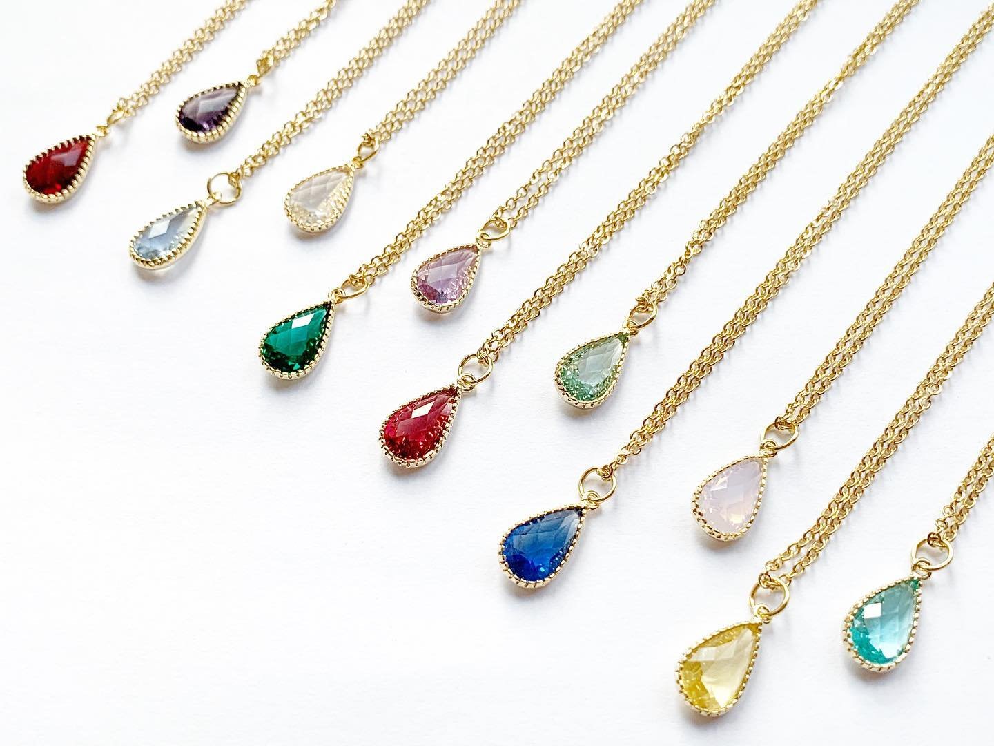 Une collection de colliers avec des pendentifs en forme de goutte, ornés de pierres précieuses colorées, disposés en ligne sur un fond blanc.