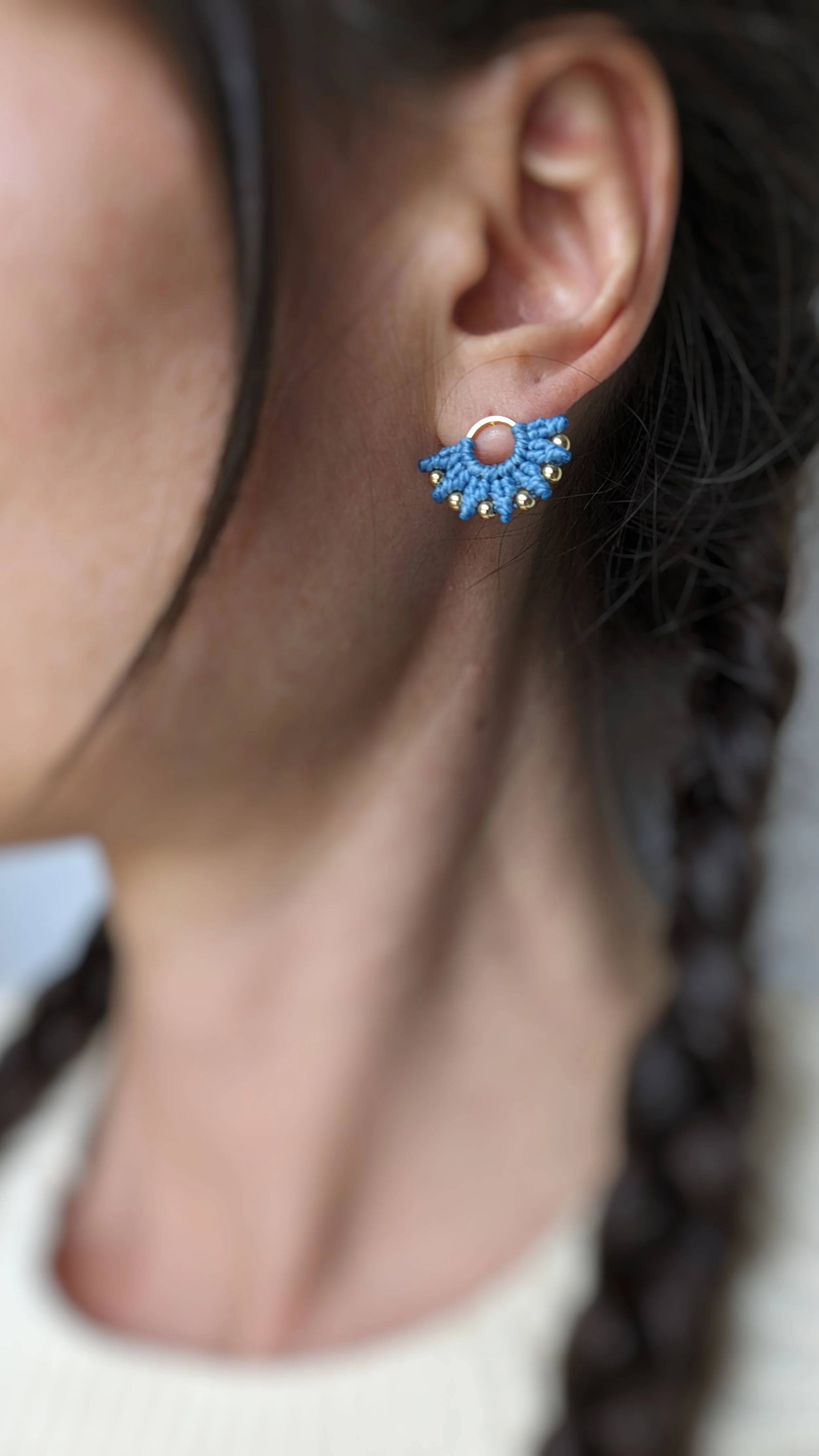 Une boucle d'oreille en forme de cercle avec un motif de macramé bleu et de petites boules dorées, portée sur l'oreille d'une femme avec des cheveux bouclés.