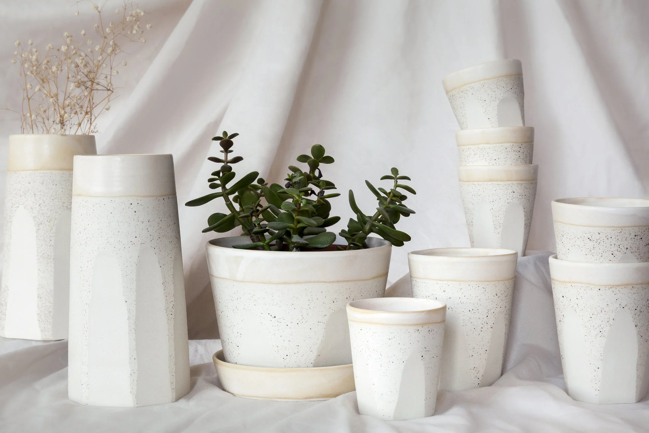 Vases en céramique blancs de différentes tailles, avec une plante succulente et des branchages séchés, disposés sur un fond de tissu blanc.