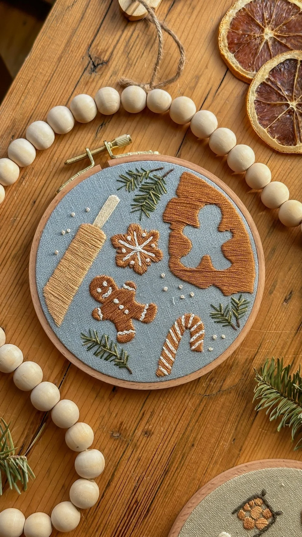 Broderie sur tissu représentant un rouleau de bonbons, un pain d'épices, un ours en pain d'épices, un flocon de neige, une branche de sapin, une boule de Noël en sucre d'orge, et une forme de feuille de trèfle, décorée avec des petites perles et des branches de sapin