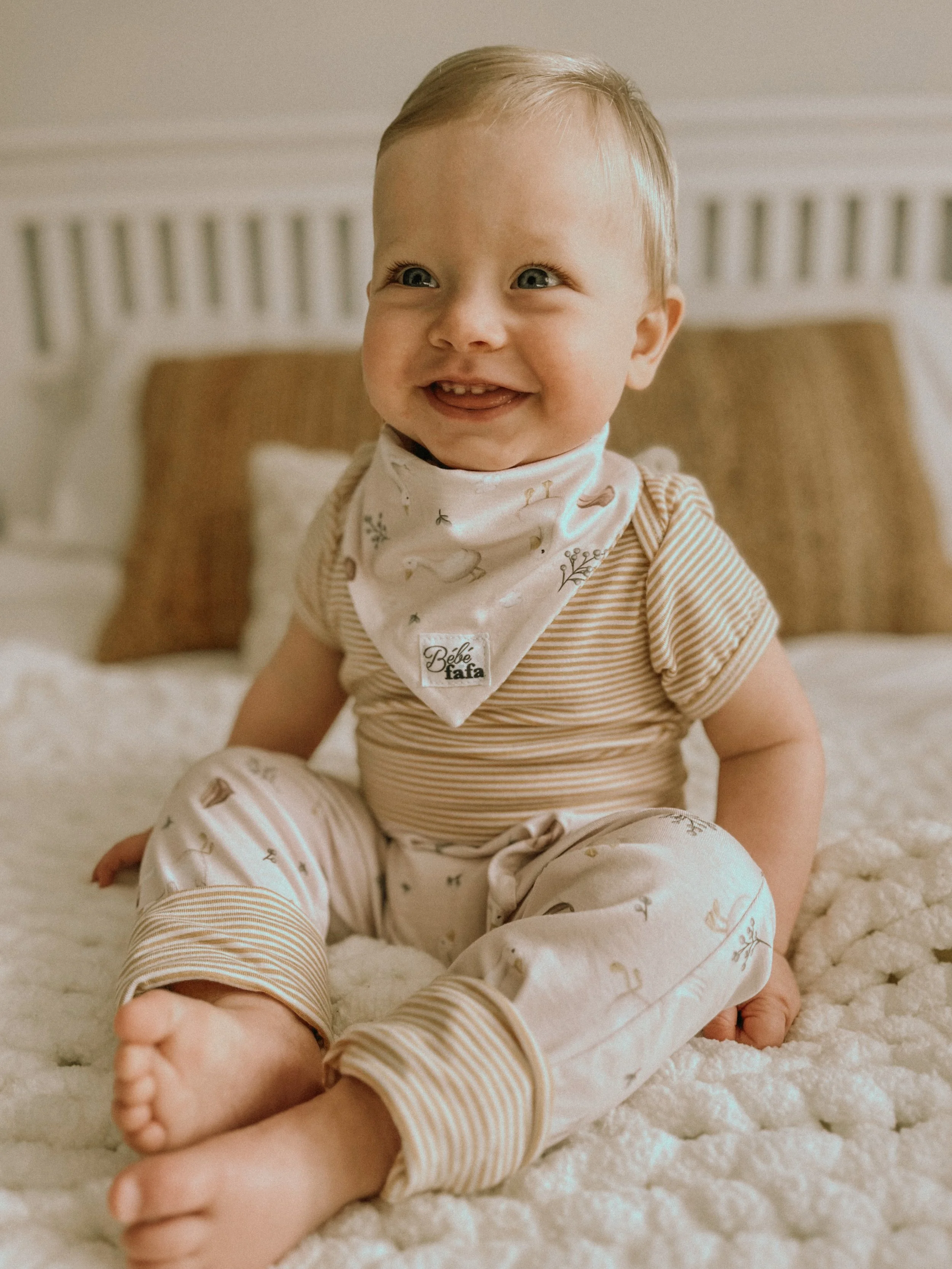 Bébé souriant assis sur un tapis beige, portant un pyjama à rayures et un bandana blanc avec des motifs d'animaux.