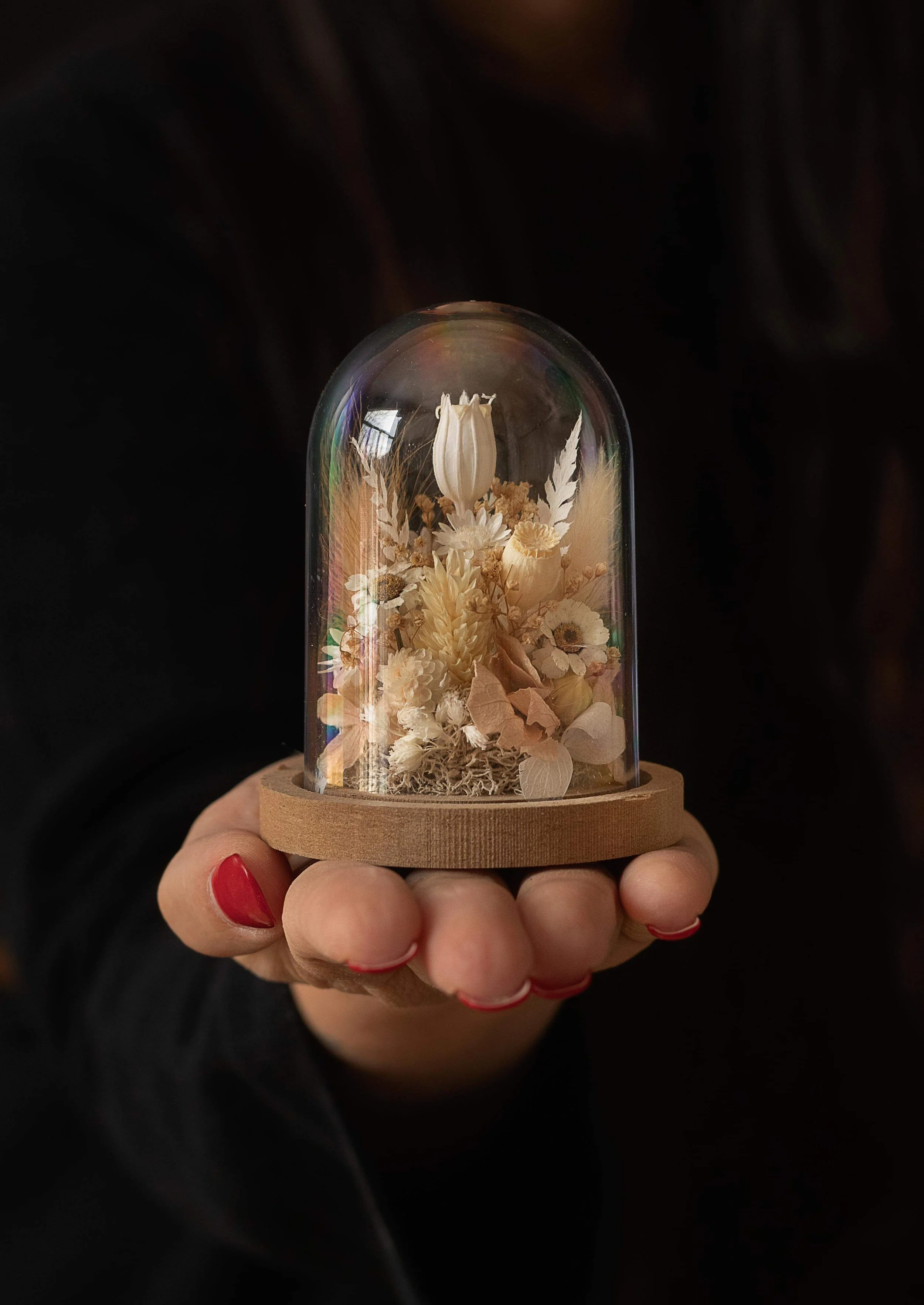 Main tenant une cage en verre contenant des fleurs séchées, avec un support en bois. La main a des ongles peints en rouge.