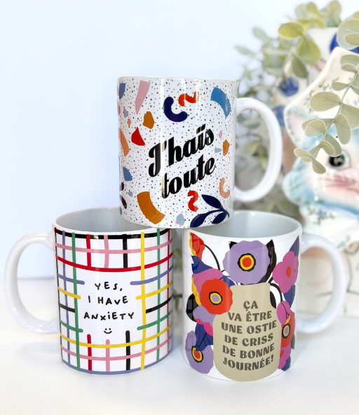 Trois mugs avec des motifs et des textes en français et en anglais, l'un avec des confettis et La phrase "J'ai touts" ou "J'ai touts" (partiellement caché), un autre avec un motif de lignes colorées et la phrase "Yes, I have anxiety", et le troisième avec un motif floral et la phrase "Ça va être une ostone de criss de bonne journée".