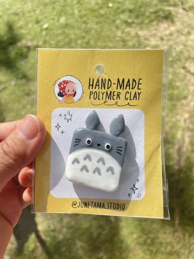 Broche en forme de créature de dessin animé, inspirée de Totoro, avec une tête grise, des oreilles pointues et un ventre blanc avec des motifs en forme de V.