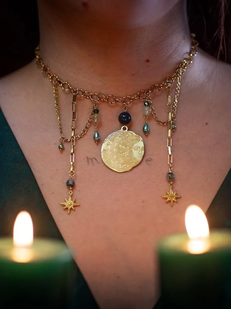 Collier en or avec pendentifs, incluant une pièce ronde avec un visage, des perles et des étoiles de mer, porté autour du cou d'une personne. Deux bougies allumées en premier plan.