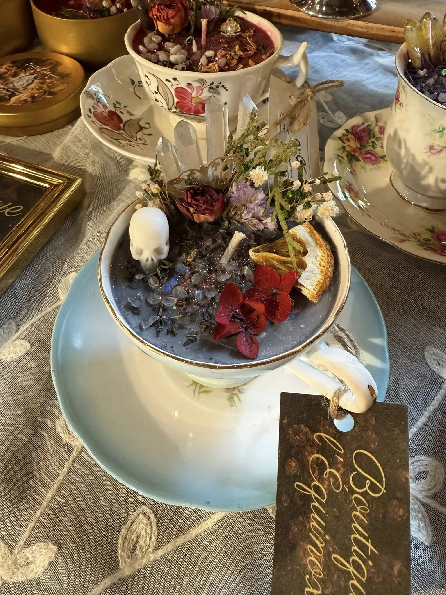 Table avec plusieurs tasses en porcelaine décorées de fleurs et contenant des fleurs séchées, une structure en cristal, des décorations florales, une petite squelettique et une tranche de orange, avec un fond de nappes en tissu et un marque-place qui dit "Bon anniversaire".