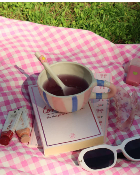 Une tasse à thé avec une cuillère, un livre, des lunettes de soleil, et divers objets décoratifs sur une couverture à carreaux roses et blancs, le tout sur de l'herbe.