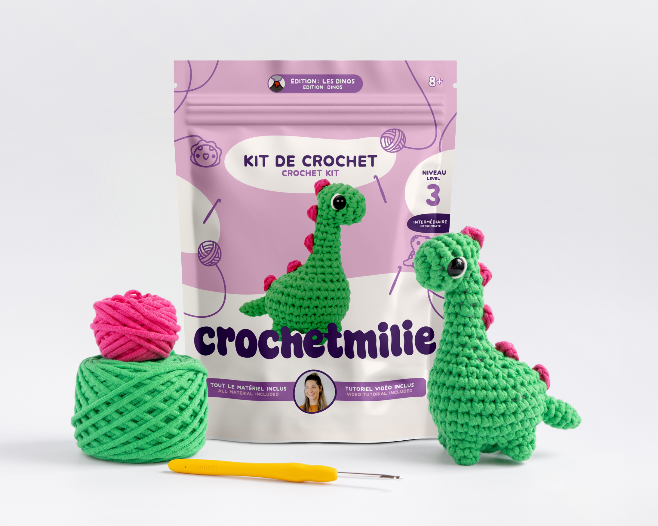 Un kit de crochet pour créer un dinosaure en laine, comprenant deux pelotes de fil colorées, un crochet jaune, et deux dinosaure en laine crochetée, avec une enceinte de présentation en arrière-plan