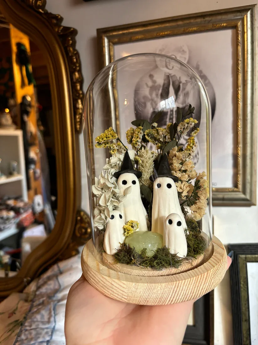 Décoration de fantômes en céramique avec des chapeaux de sorcière, entourée de fleurs séchées, sous une coupelle en verre. Contexte intérieur avec miroir et cadre doré.