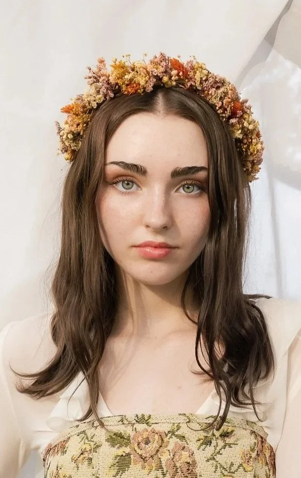 Une jeune femme avec des yeux verts porte une couronne de fleurs colorées sur la tête et un haut à motifs floraux.