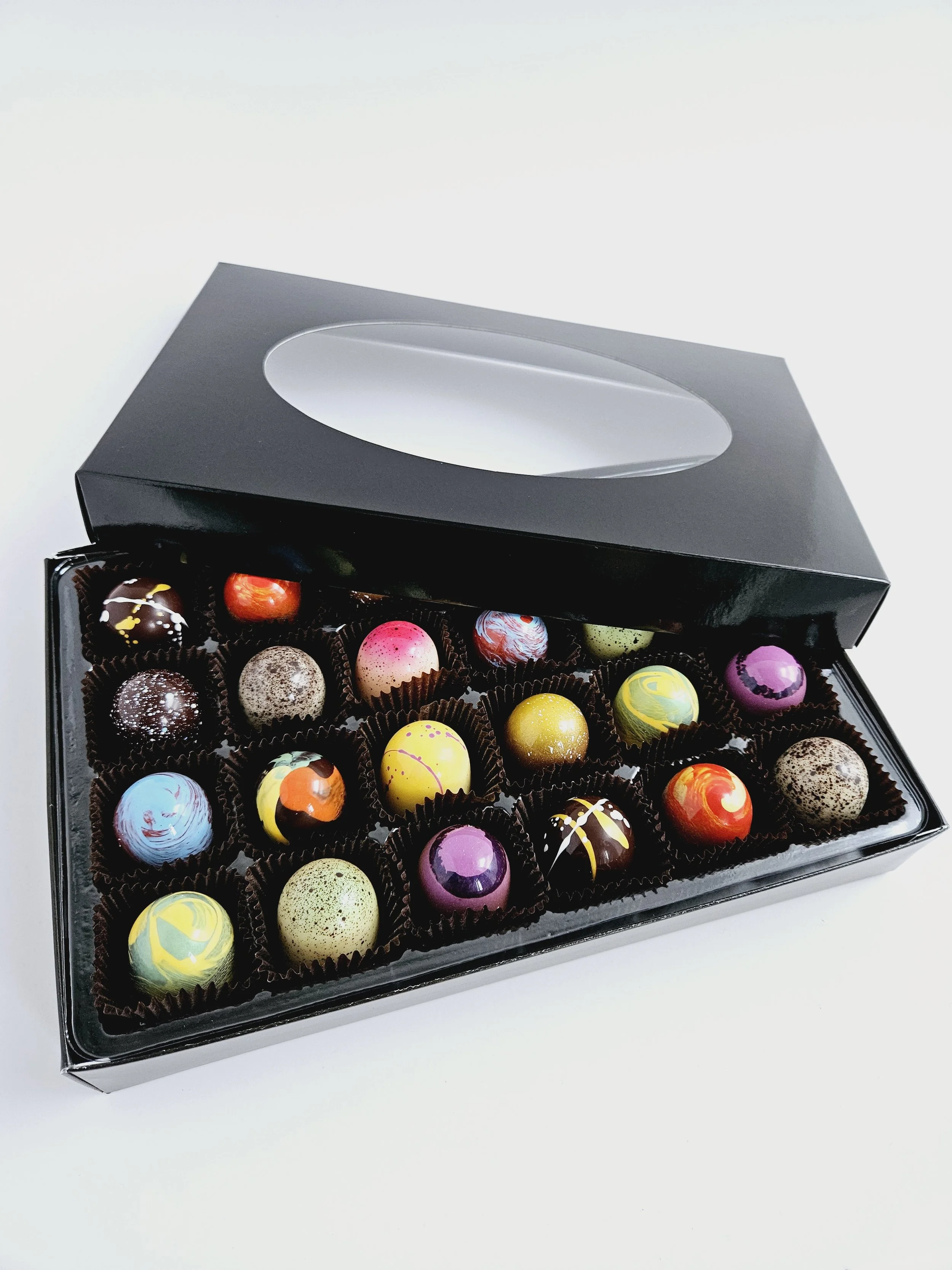Une boîte de chocolats artistiquement décorés, avec une variété de couleurs et de motifs, dans une boîte en plastique avec un couvercle transparent.