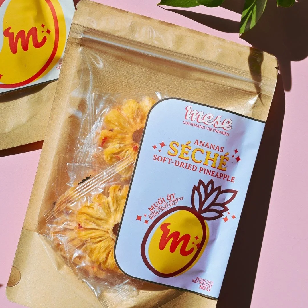 Paquet de ananas séché à base d'ananas doux et salé, en emballage transparent avec un label blanc, jaune et rouge, posé sur une surface rose, avec une plante verte à côté.
