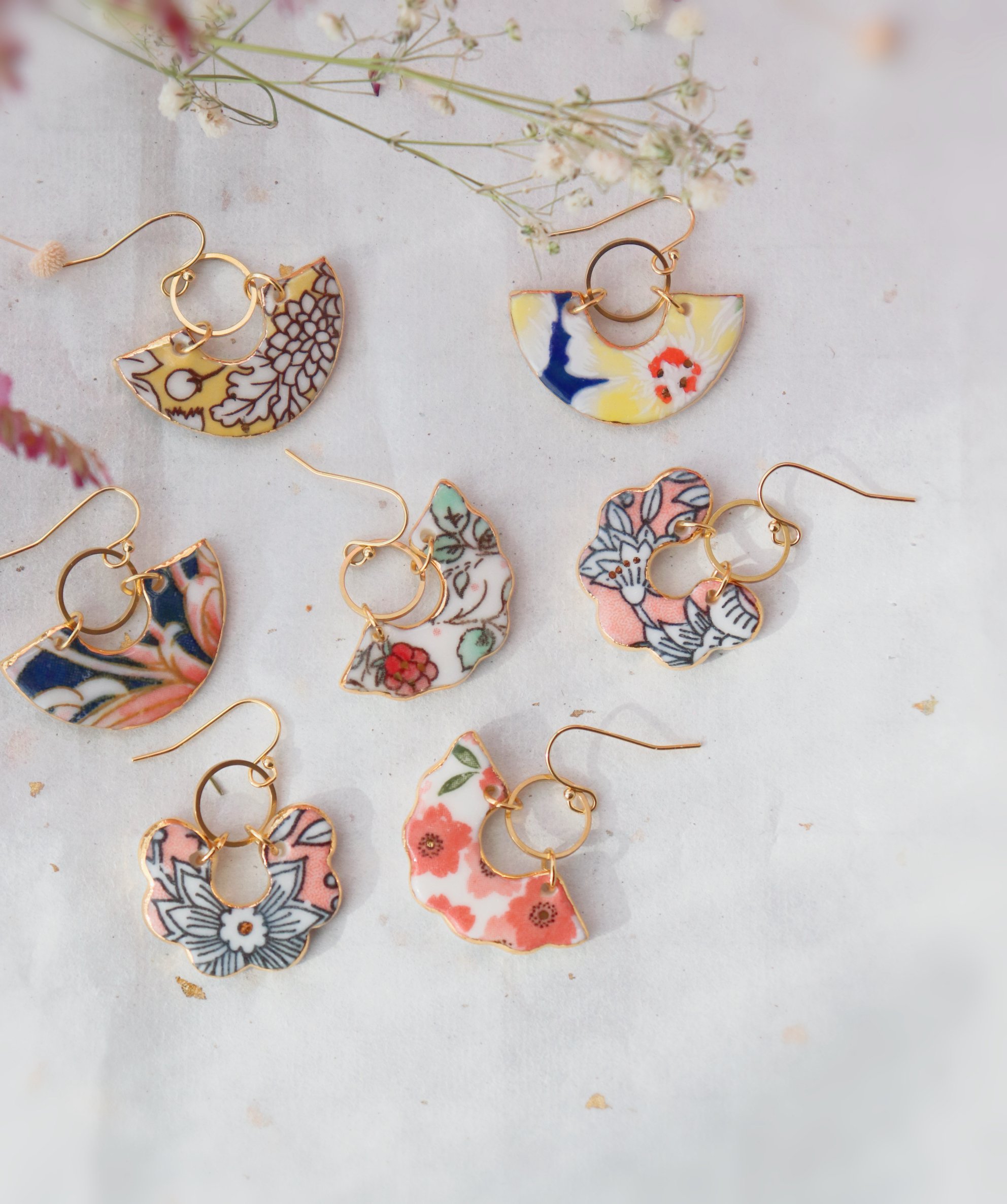 Une paire de boucles d'oreilles en morceau de céramique peint avec des motifs floraux et colorés, posées sur une surface claire avec des fleurs séchées en arrière-plan.