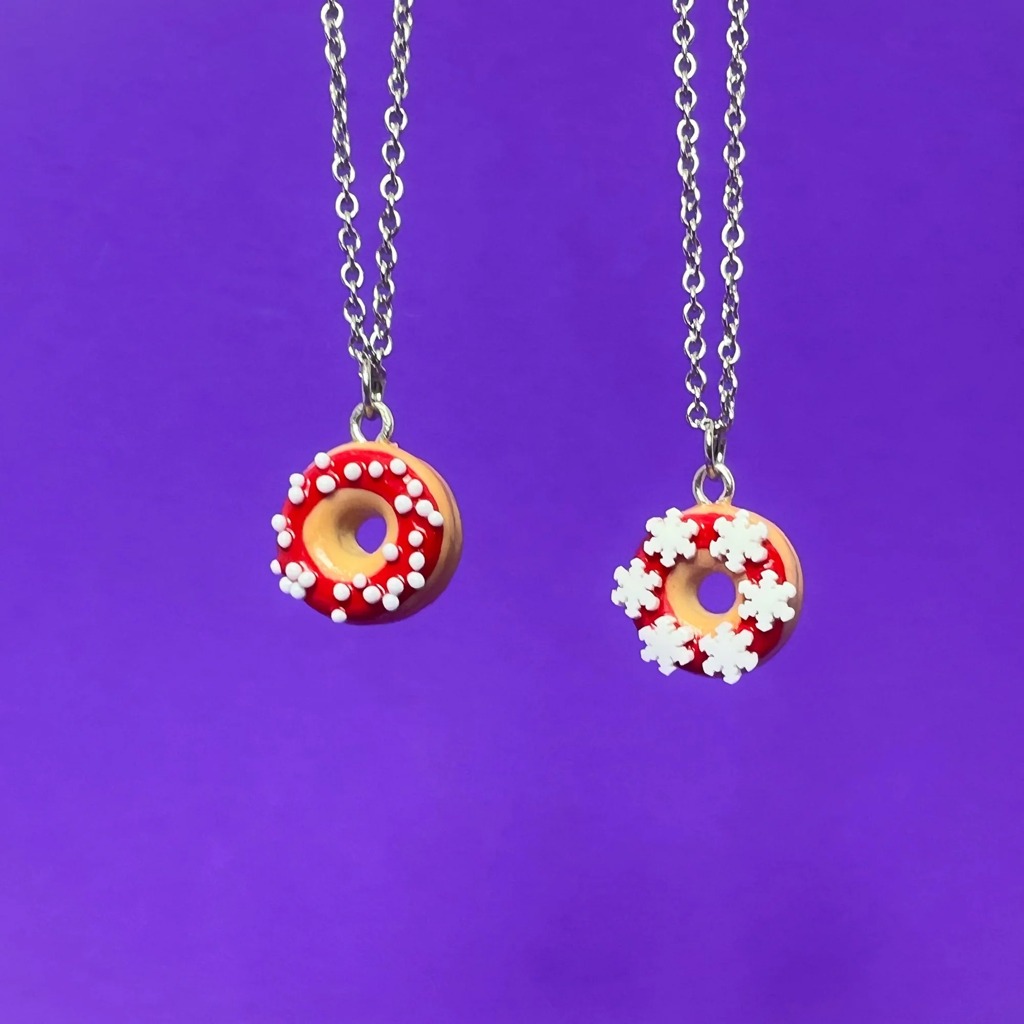Des colliers à pendentif donuts avec glaçage rouge et décoration de petites fleurs blanches, suspendus contre un fond violet.
