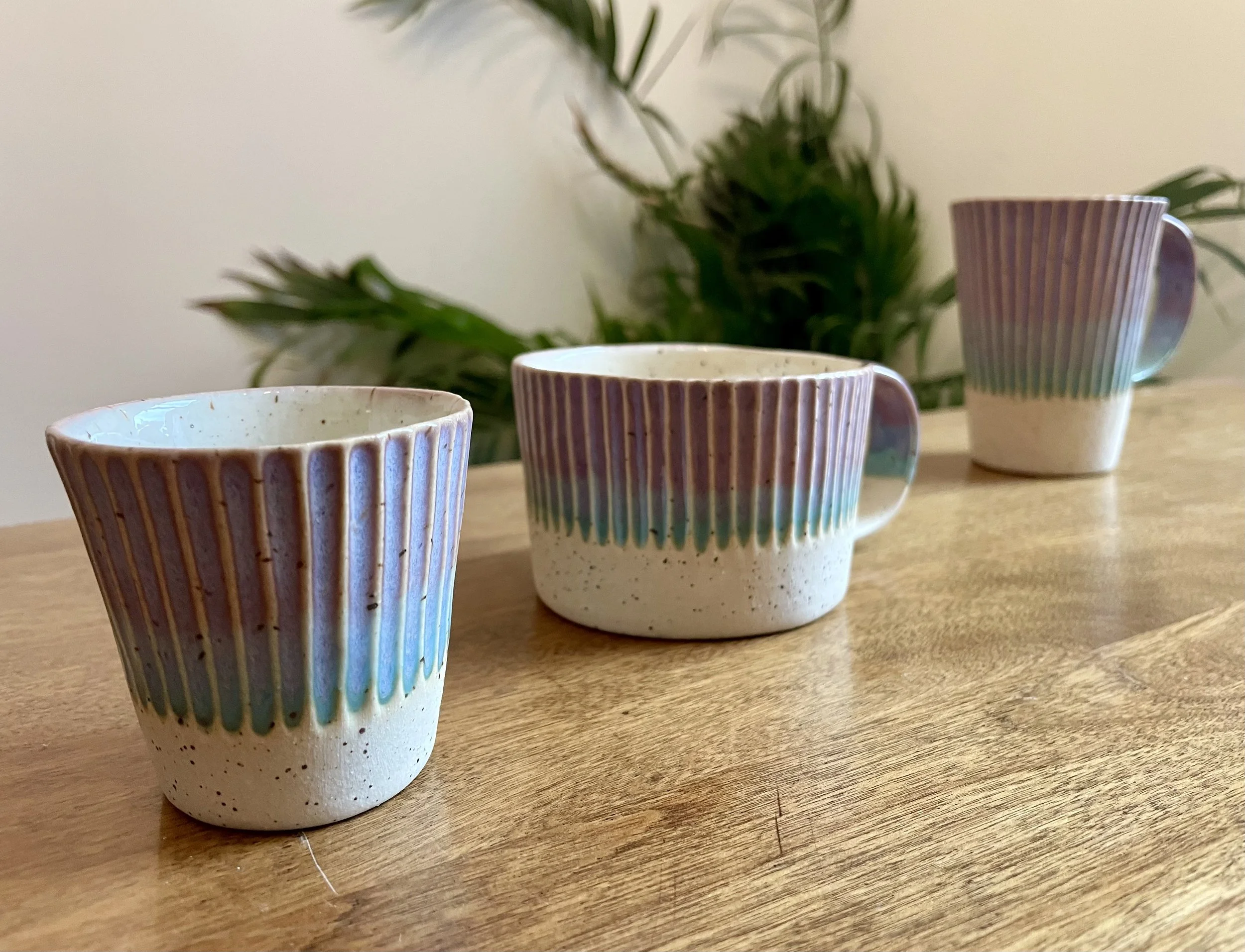 Trois tasses en céramique avec un motif rayé pastel, posées sur une surface en bois, avec une plante verte en arrière-plan.