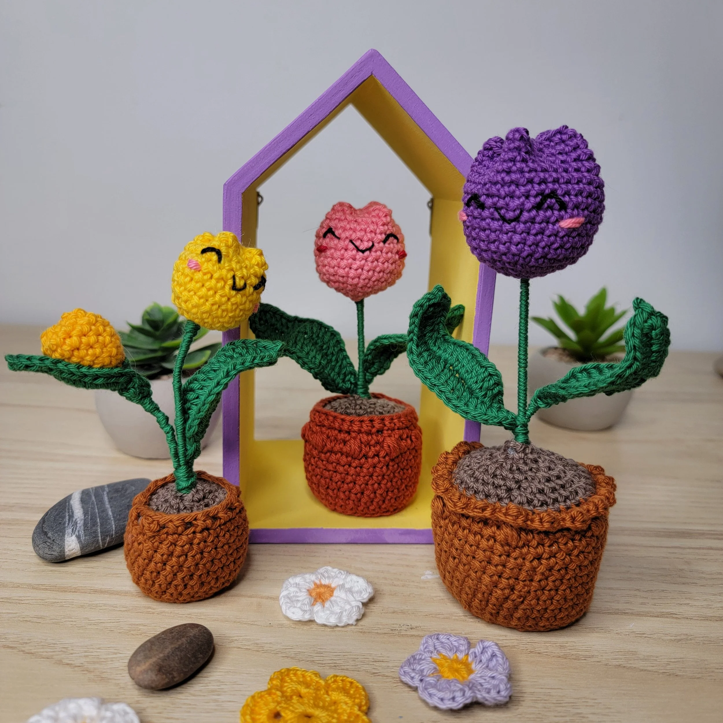 Une décoration de fleurs en laine tricotée, avec différentes fleurs de couleurs jaune, rose, violet et orange, placées sur une table en bois. Certaines fleurs ont des visages souriants et des feuilles vertes en laine.