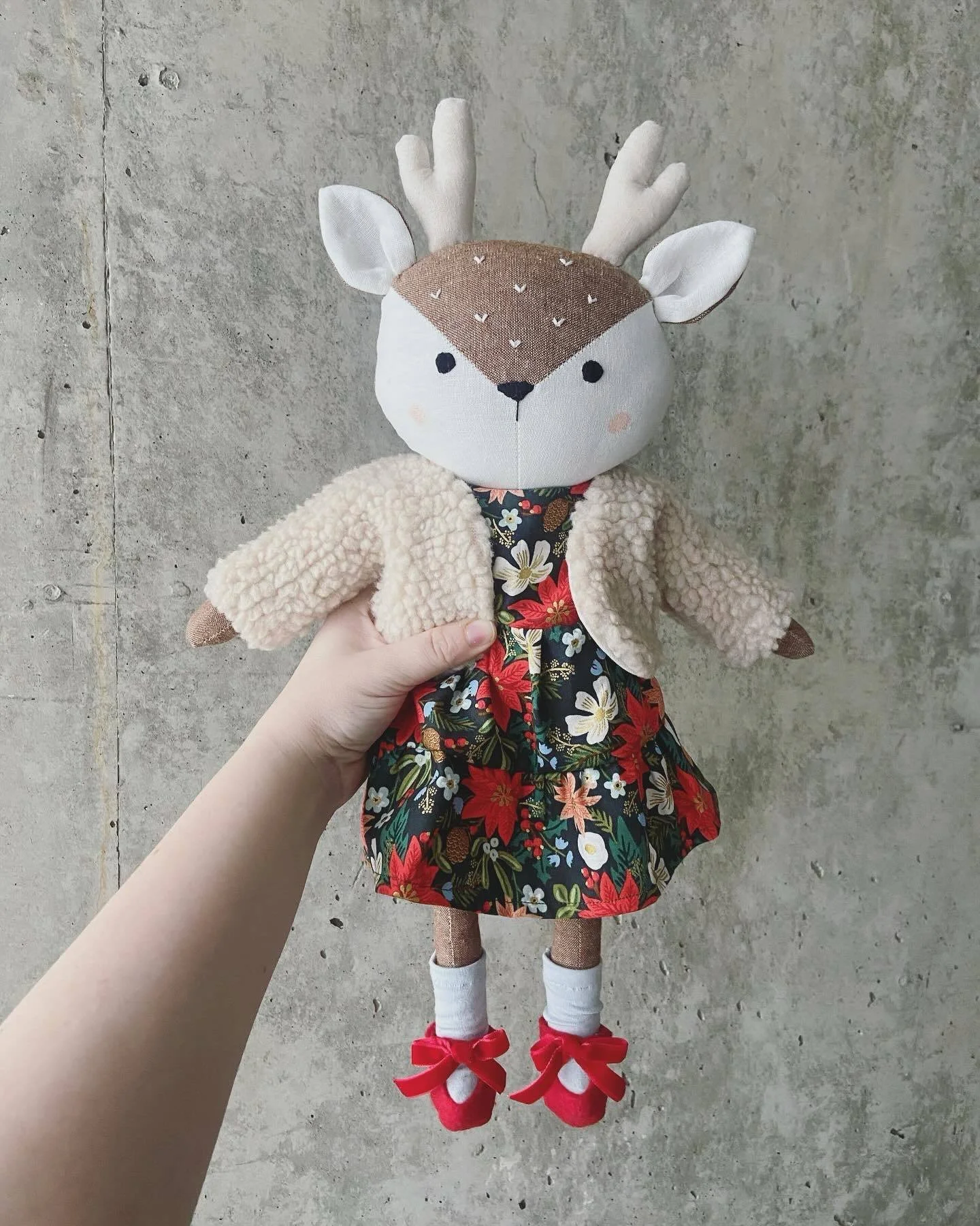 Poupée en forme de cerf avec une tête en tissu beige, des oreilles blanches, et des bois en tissu beige. La poupée porte une robe à motifs floraux, un manteau en tissu peluche beige, des chaussettes blanches avec des chaussures rouges à nœuds.