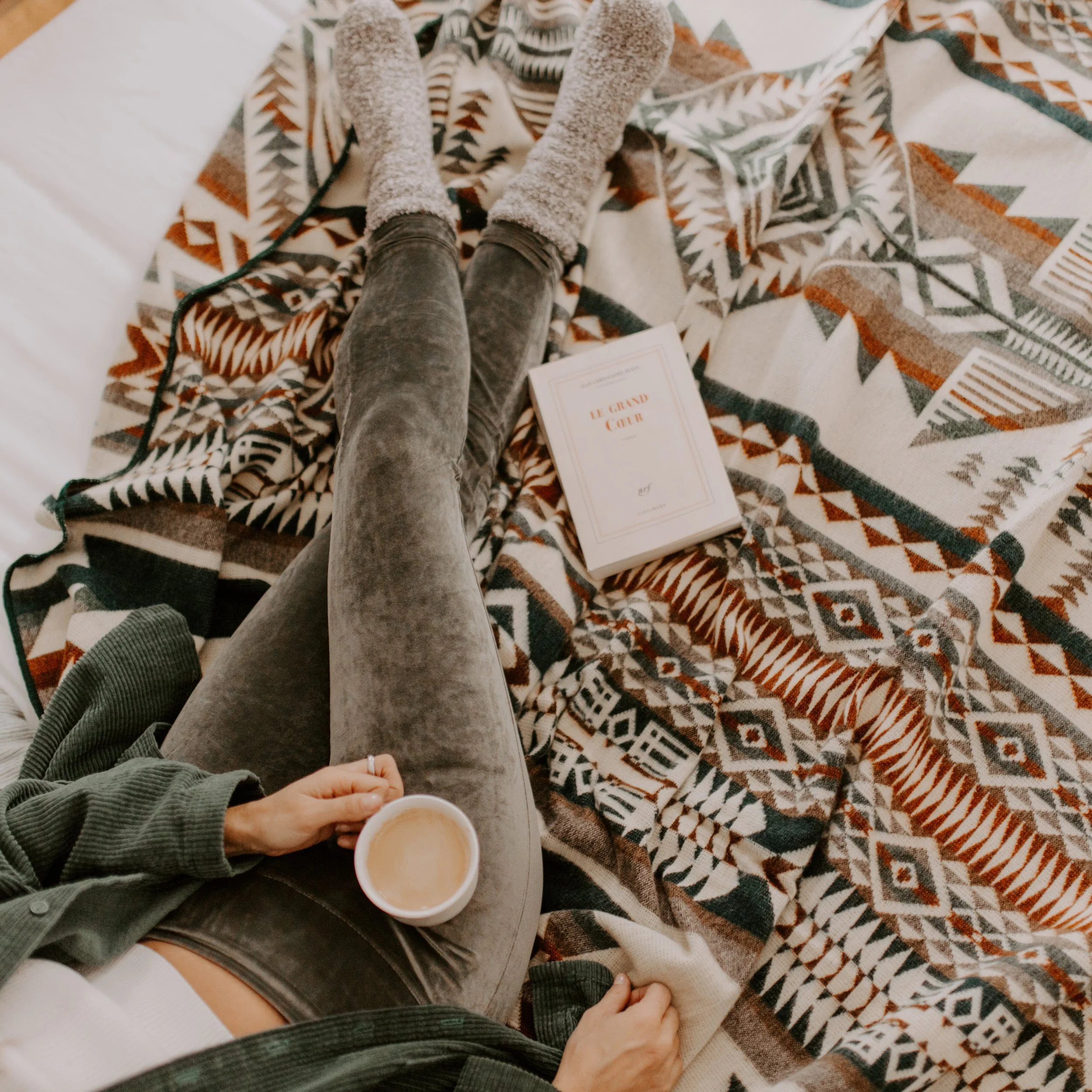 Une personne tient une tasse de café chaud dans une main, assise sur un canapé avec une couverture à motif géométrique. Il y a deux autres tasses de café ou de thé sur la table, ainsi qu'un livre intitulé 'Le Grand Oui'. La personne porte un pull vert.