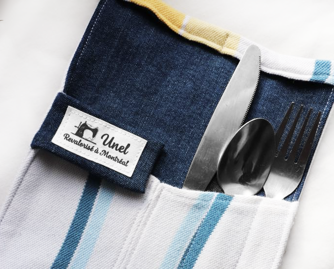 Une poche de serviette en tissu avec couvercle, contenant une fourchette, une cuillère et un couteau en métal, avec un tag indiquant "Unel" et "Revalorisé à Montréal".