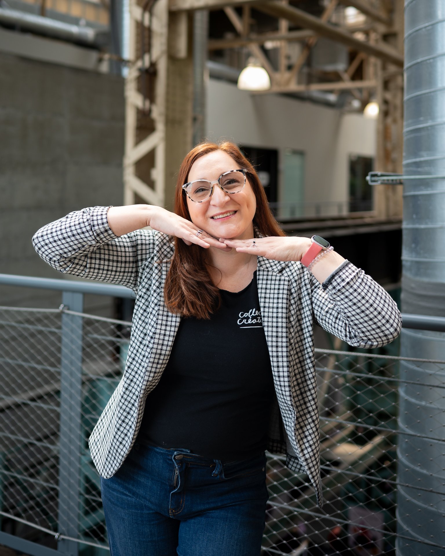 Une femme avec des lunettes, portant une veste à carreaux et un t-shirt noir, pose avec un sourire, le visage détendu, dans un environnement urbain avec des structures en bois et des éléments métalliques.