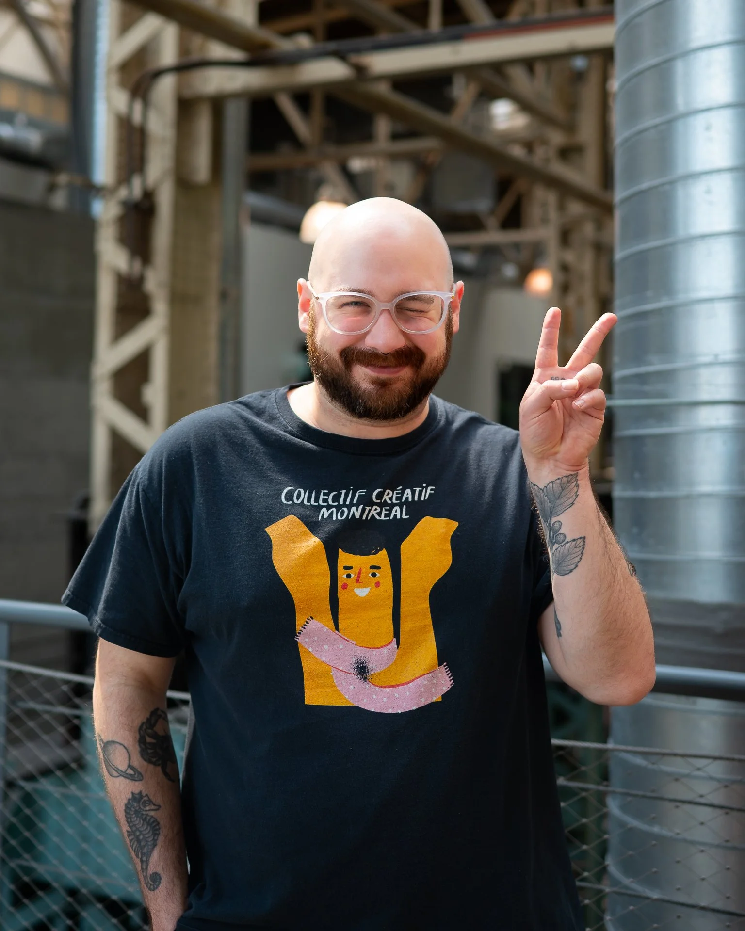 Un homme chauve avec une barbe porte un t-shirt noir avec un dessin d'une femme portant un bracelet en forme de serpent. Il porte des lunettes transparentes et fait un signe de paix avec la main droite. Il sourit dans un environnement industriel avec des tuyaux en métal et une structure en bois en arrière-plan.