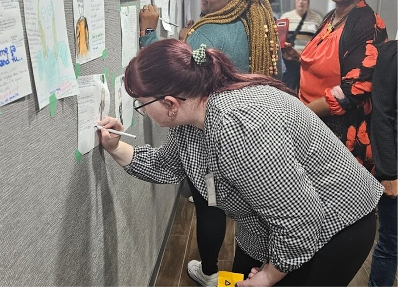 Woman with red hair and glasses writes on paper attached to a gray bulletin board at an event with other people in the background.