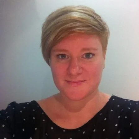 A woman with short blonde hair wearing a black top with small white dots, smiling against a plain light-colored wall background.