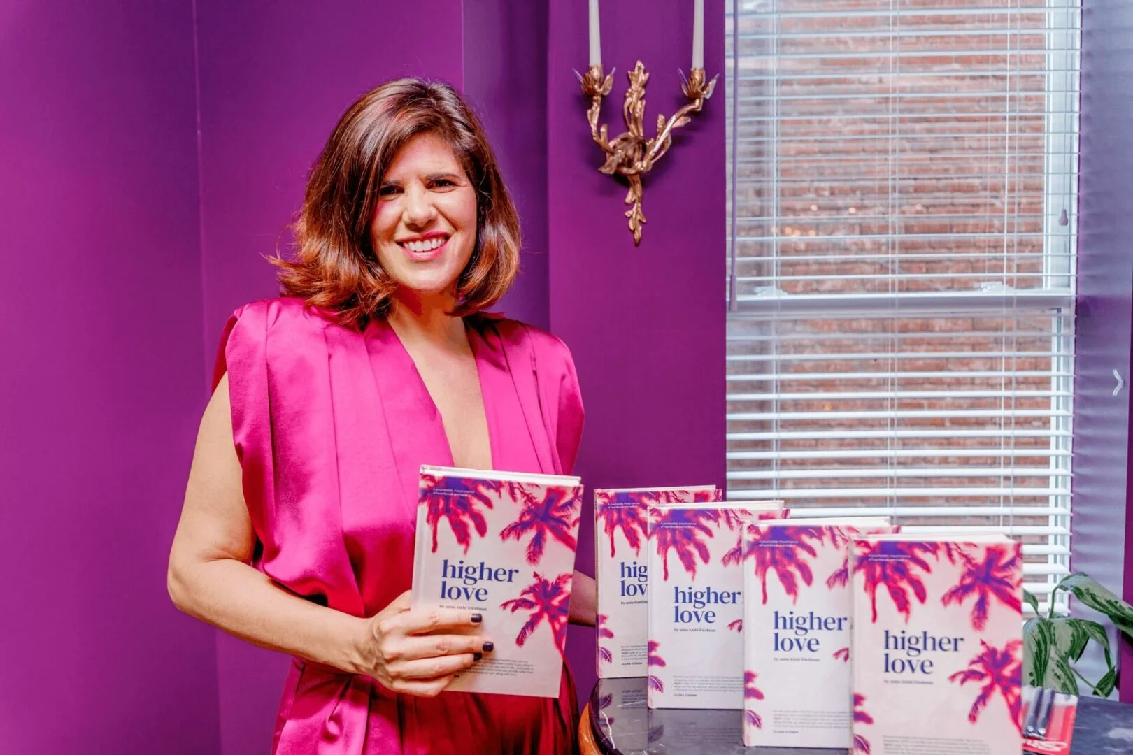 A woman in a pink satin top holding a book titled 'Higher Love' with palm tree illustrations on the cover, standing by a purple wall and a window with blinds, next to several copies of the same book on a table.