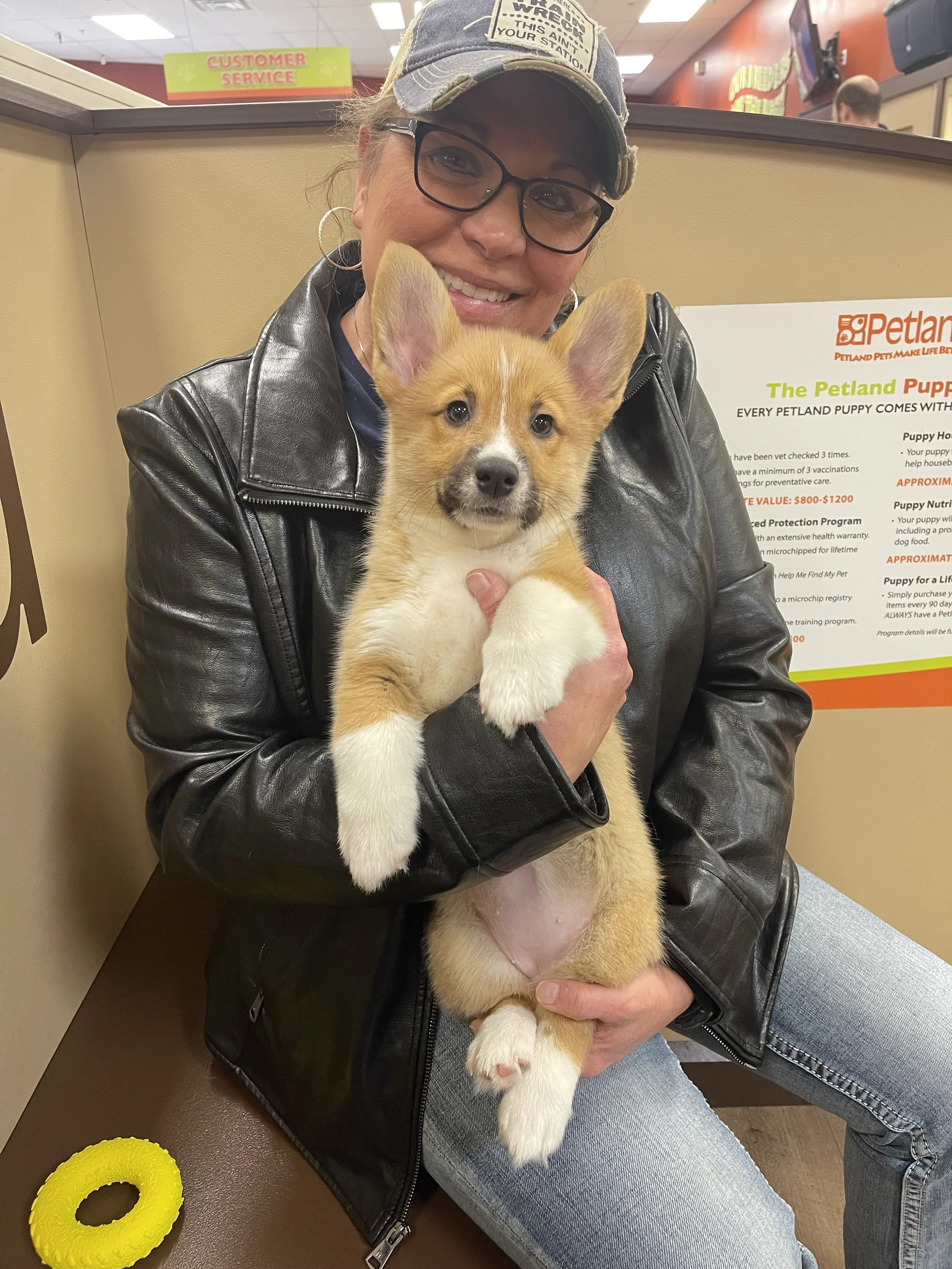 A woman with glasses, hoop earrings, a baseball cap, and a black leather jacket is sitting down, holding a cute puppy with large ears and a light brown and white coat in her lap. The setting appears to be a pet store or veterinary clinic, with informational posters and signs in the background.