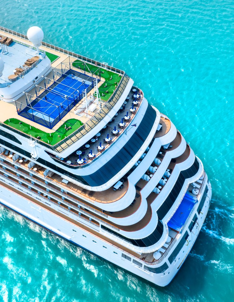Aerial view of a large cruise ship with multiple decks, pools, and recreational areas sailing on turquoise water.