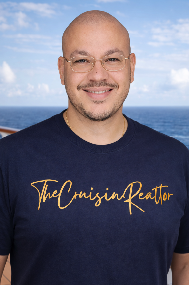 A smiling man wearing glasses and a navy blue t-shirt with gold script that says "The Cruisin Realtor" stands outdoors with a ship's deck and ocean in the background.