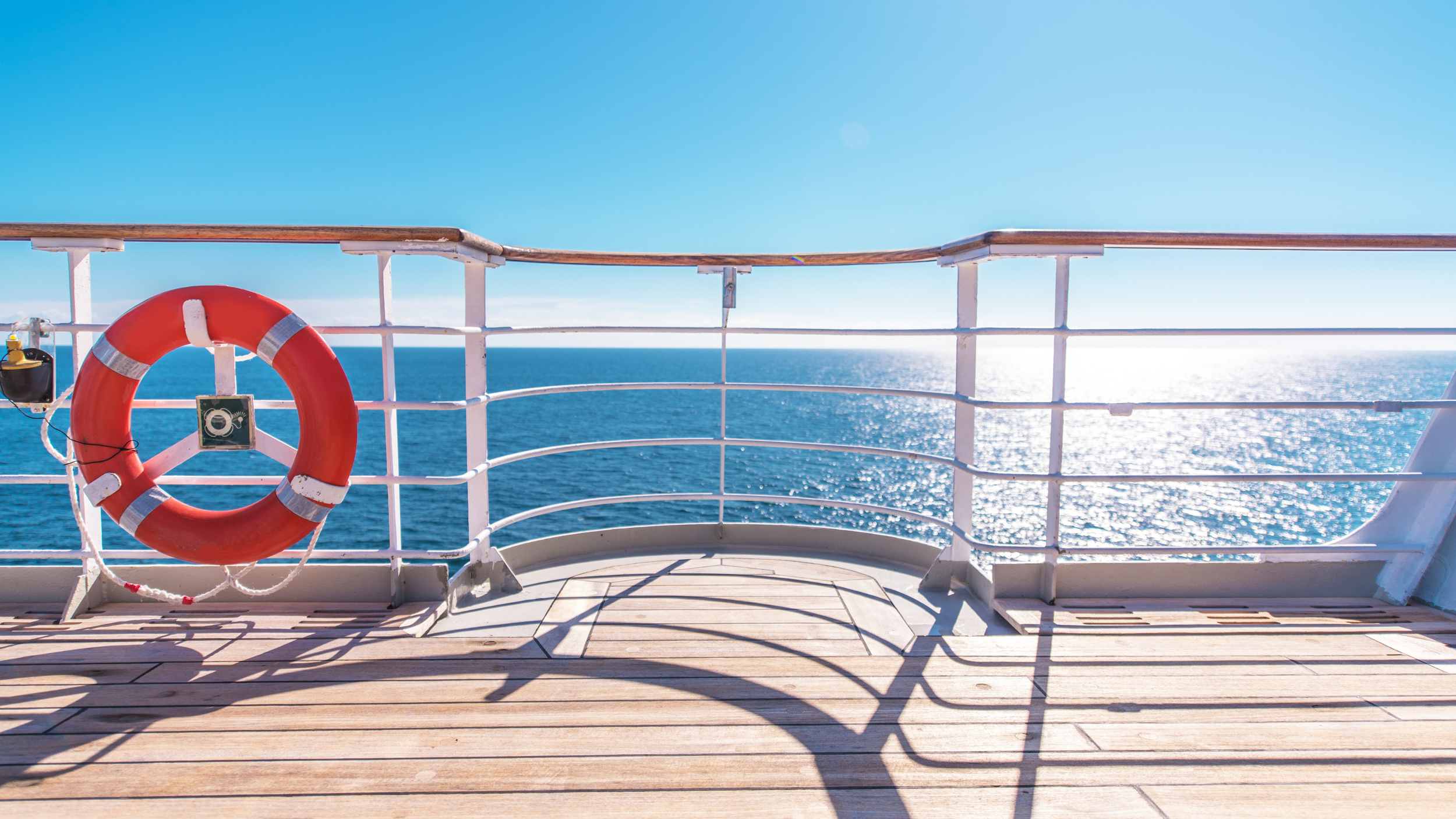 View from a ship's deck showing a white railing, an orange life preserver, and the ocean with sunlight reflecting off the water, under a clear blue sky.