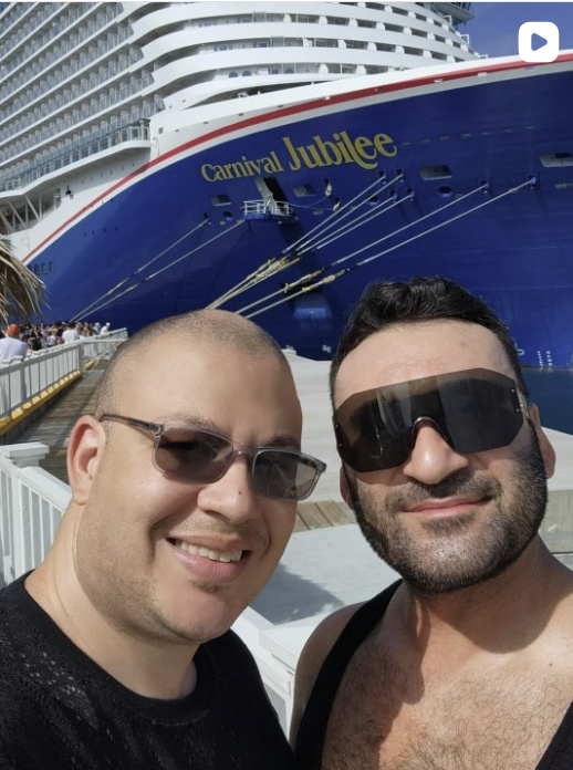 Two men taking a selfie in front of the cruise ship Carnival Jubilee, docked at a port with a crowd of people in the background.