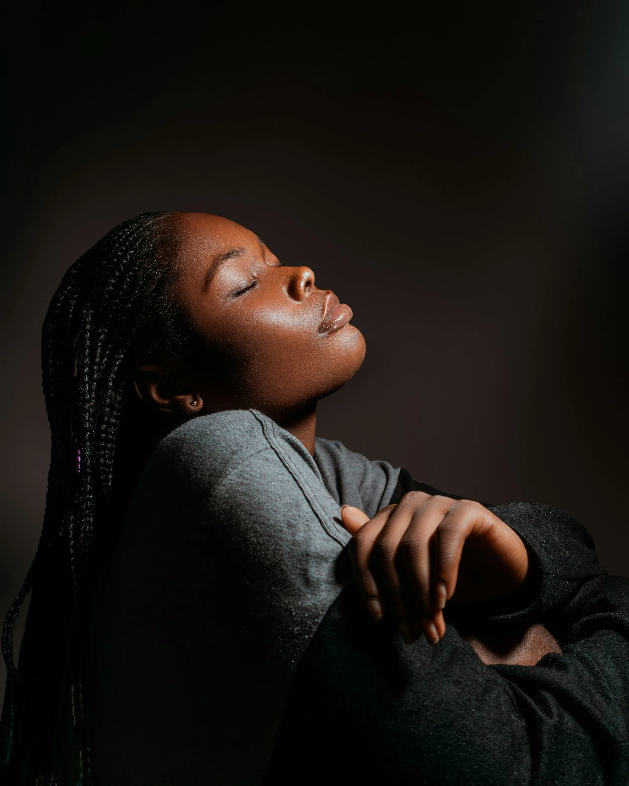A woman with closed eyes and braided hair, appearing relaxed with hands crossed over her chest, against a dark background.