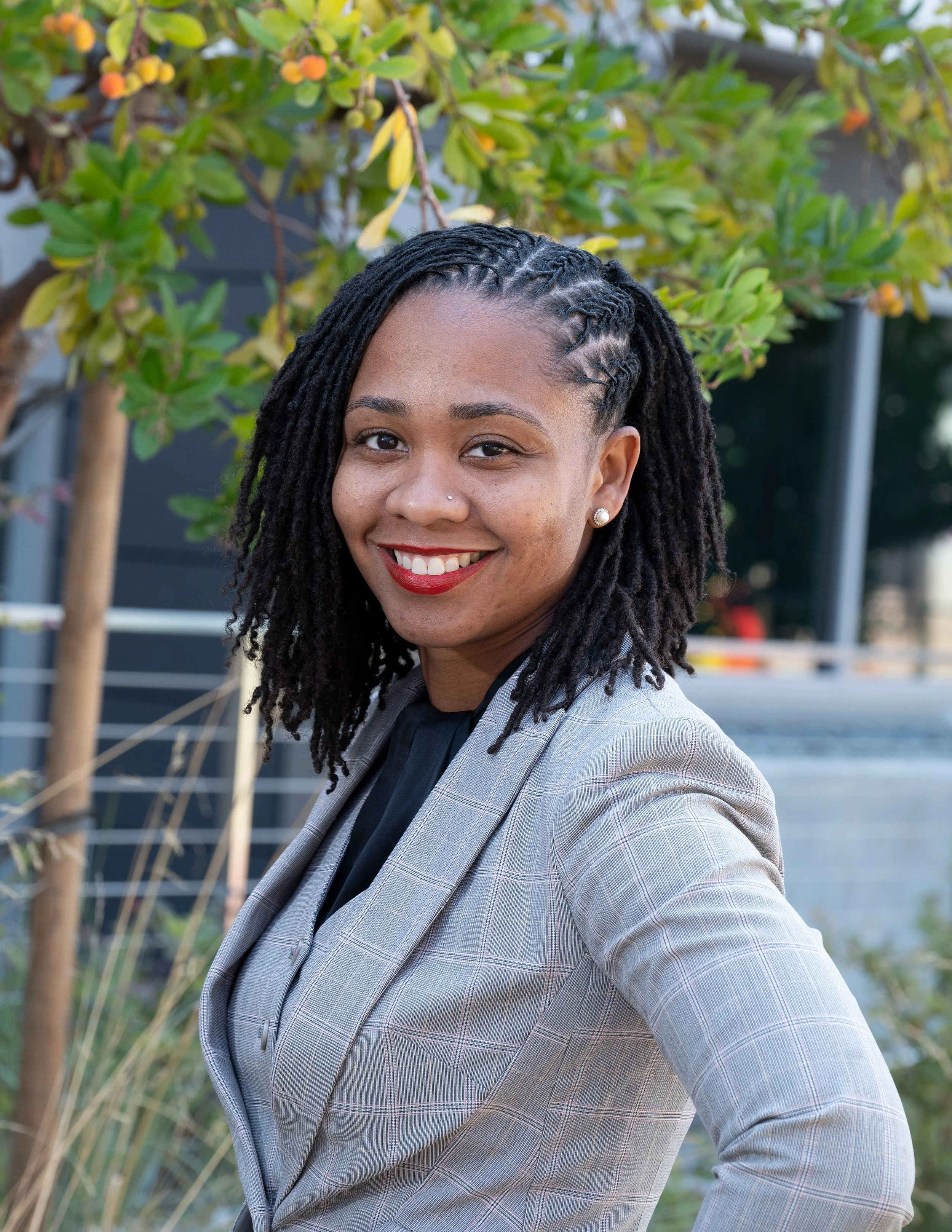 A smiling woman with long dreadlocks, wearing a plaid blazer and pearl earrings, standing outdoors near green bushes.