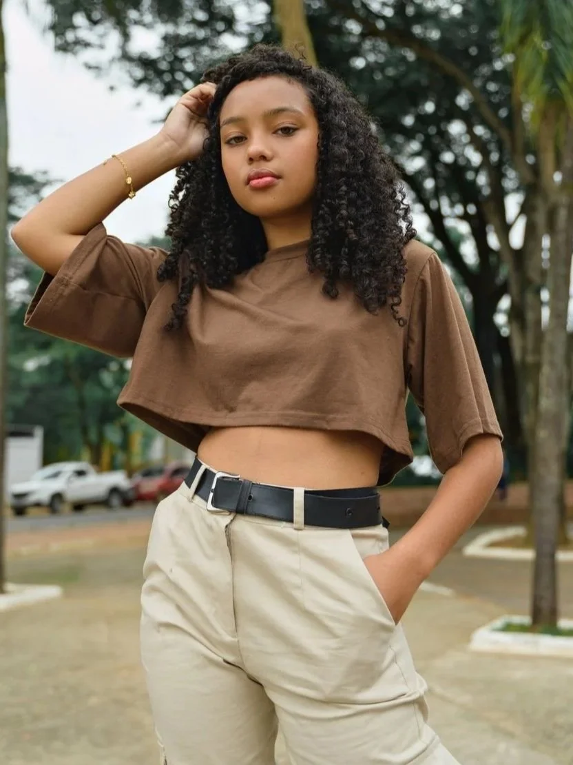 Young woman with curly hair and a confident expression, standing outdoors in a park-like setting with trees and parked cars in the background, wearing a brown crop top and beige pants.