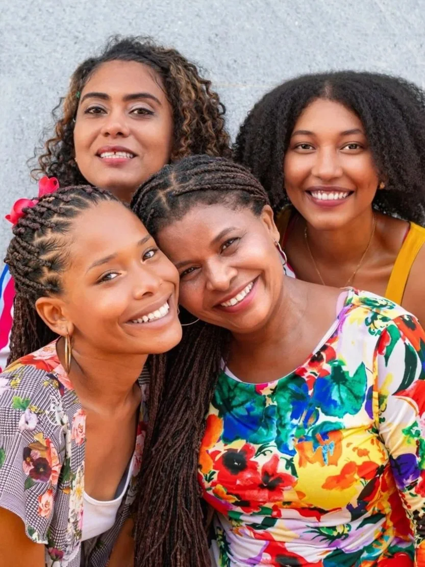 Four women smiling and standing close together outdoors against a light-colored wall.