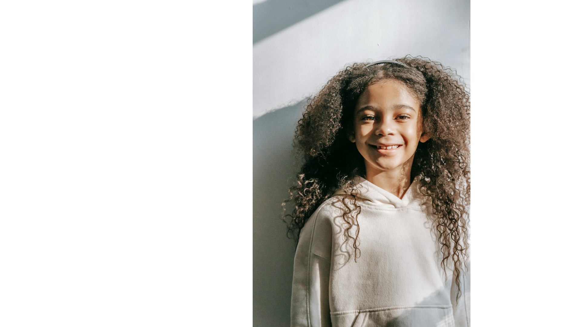 Close-up of a young girl with long curly hair, wearing a light-colored hoodie, smiling, standing against a white wall with sunlight casting a shadow.