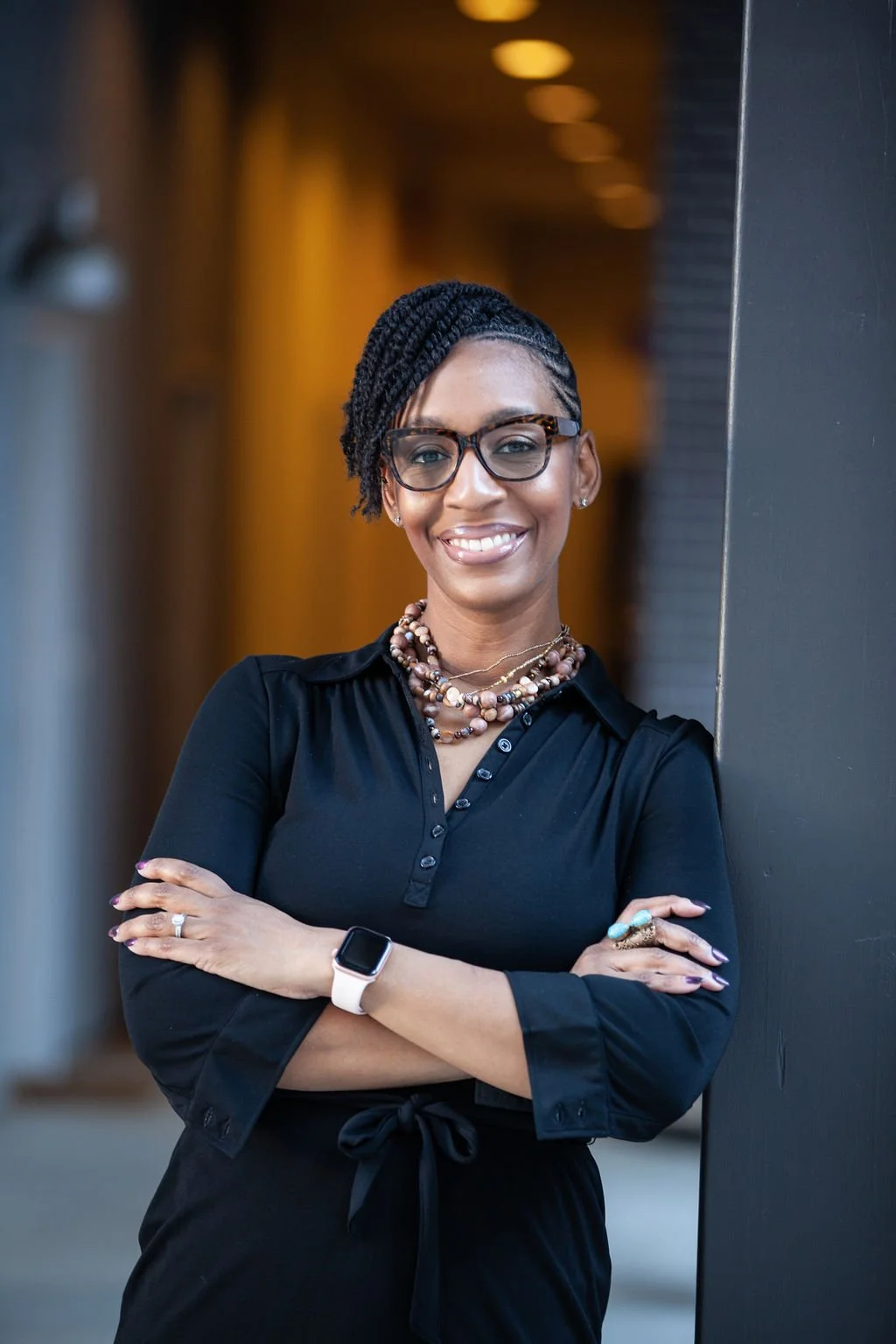 A confident woman with glasses, braided hair, and a bright smile, standing with arms crossed against a dark wall in an indoor setting.