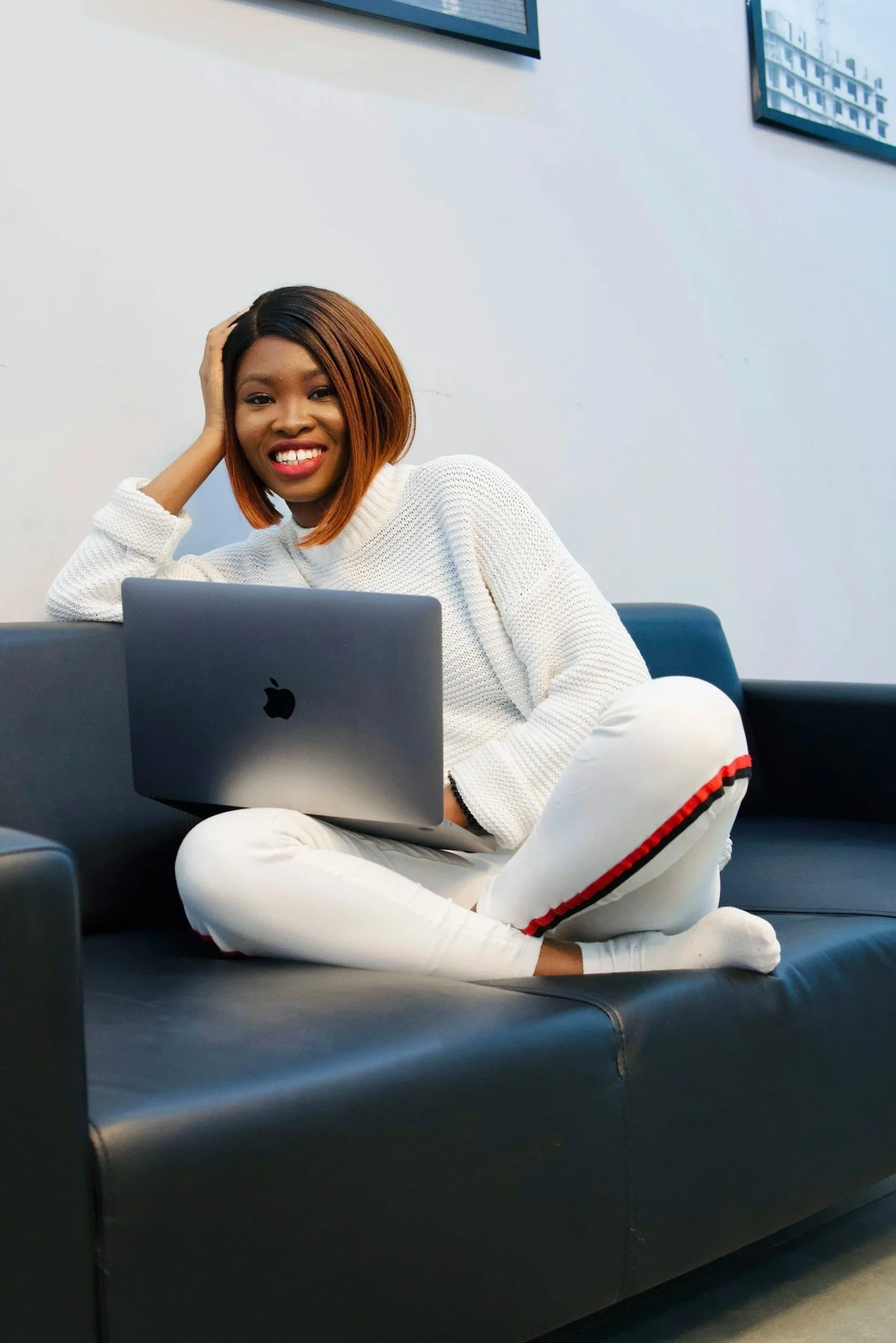 A woman with shoulder-length brown hair, wearing a white sweater, sitting on a black leather couch in a modern office setting. She is smiling and resting her head on her left hand. In front of her is an open laptop, and there are framed abstract artwork on the white wall behind her.