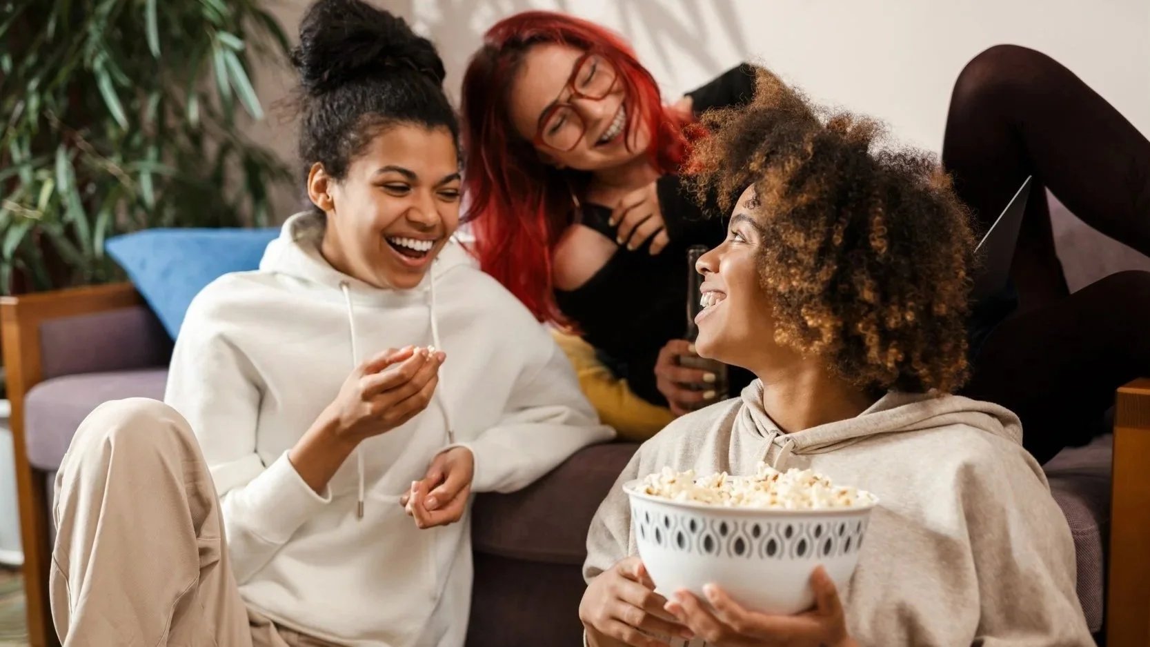 Four women enjoying a movie night at home, with one holding a bowl of popcorn and all laughing together.
