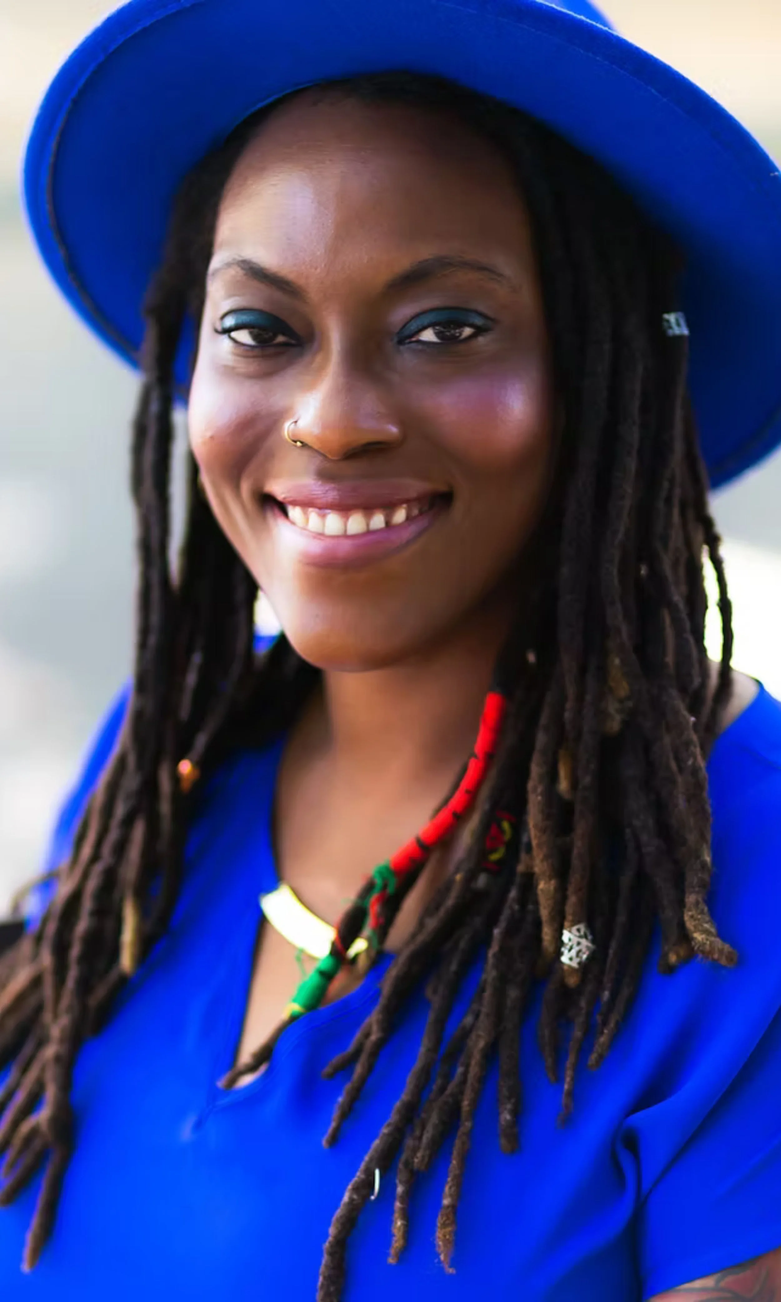 A smiling woman with long dreadlocks wearing a wide-brimmed blue hat and matching blue top, accessorized with a colorful beaded necklace and a gold nose ring.