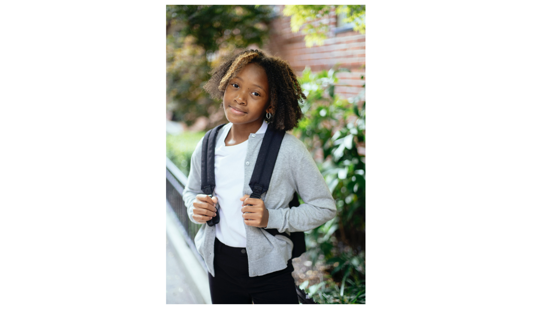 A young girl with curly hair, wearing a gray cardigan, white shirt, and black pants, standing outdoors with a backpack, smiling gently at the camera.