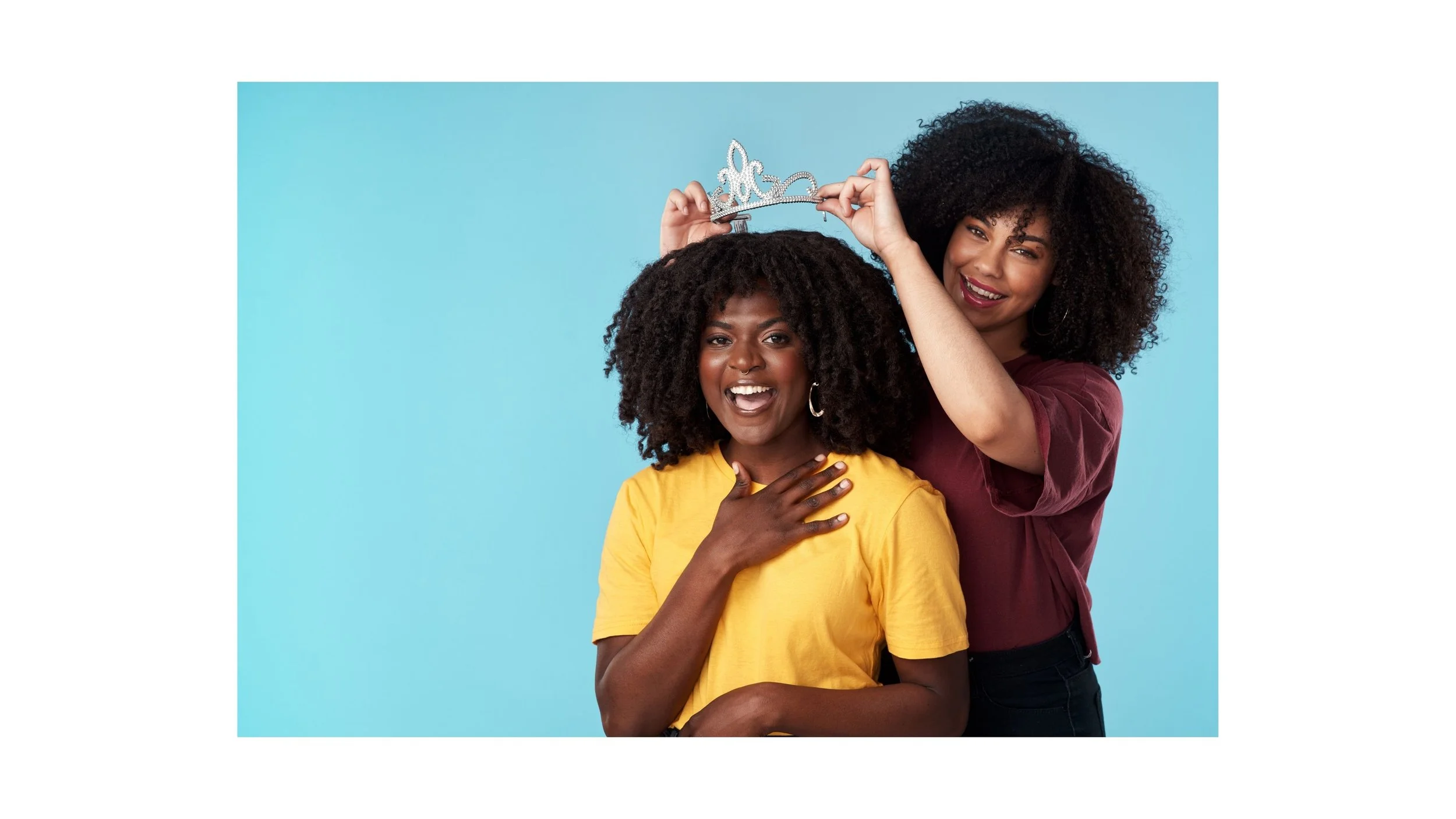 Two women with curly hair smiling, one wearing a yellow shirt and the other in a burgundy shirt, holding a tiara together against a light blue background.